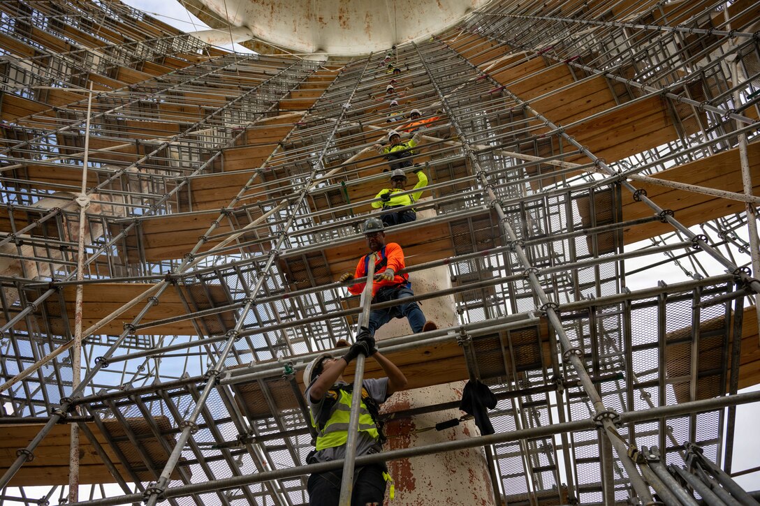 Four men stand one above another on different levels of scaffolding and they pass a metal pipe up to one another.