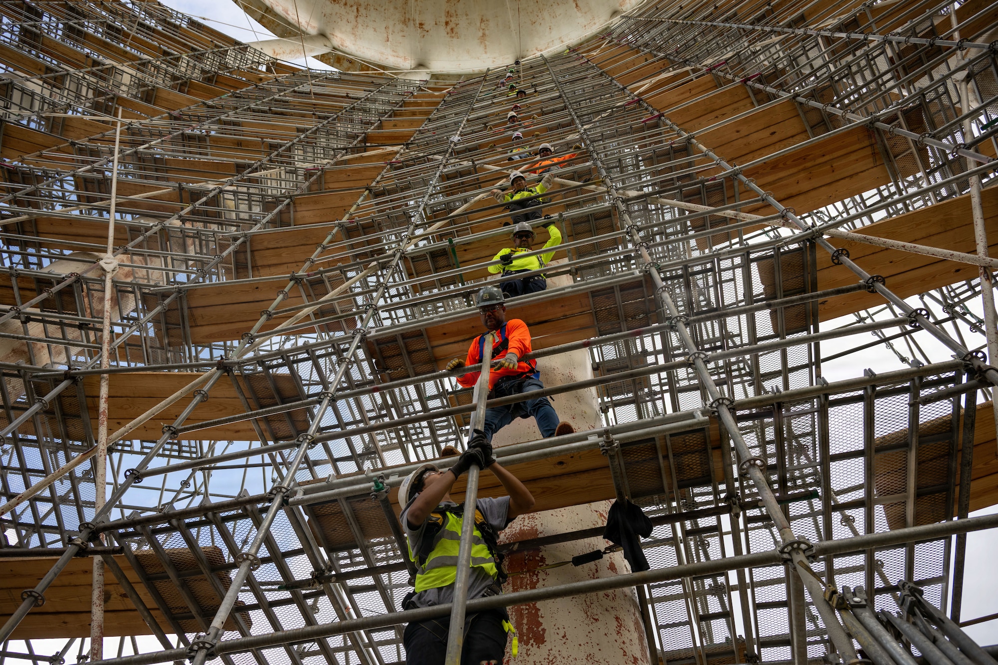 Four men stand one above another on different levels of scaffolding and they pass a metal pipe up to one another.
