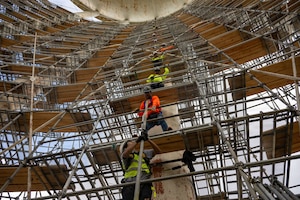 Four men stand one above another on different levels of scaffolding and they pass a metal pipe up to one another.
