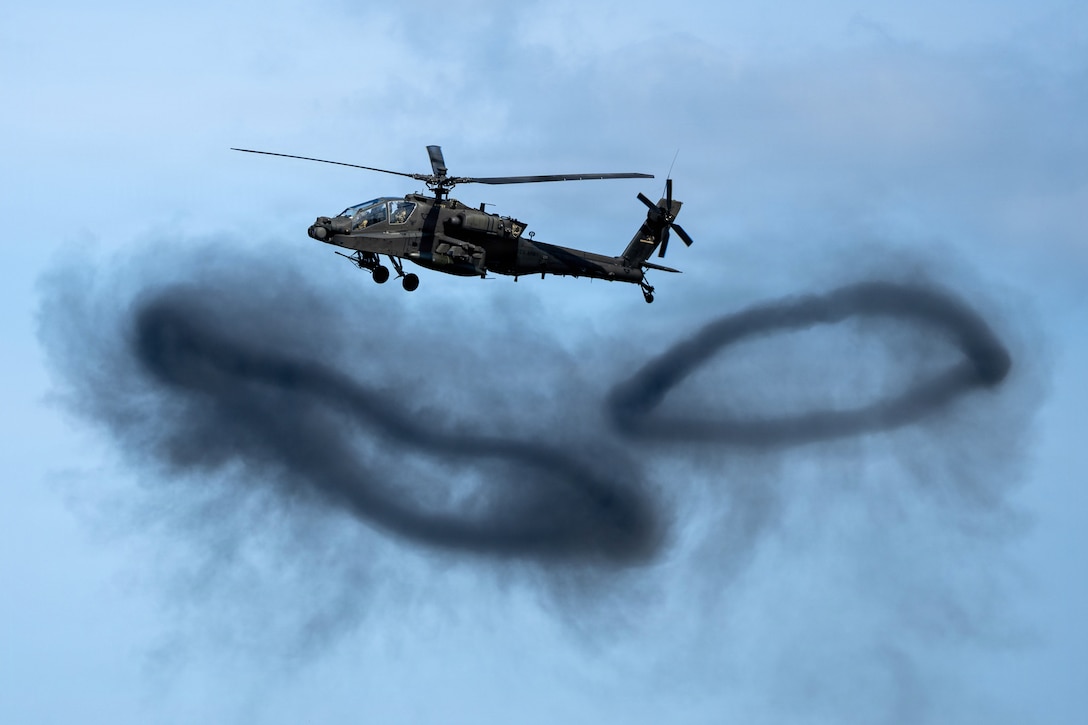 A military helicopter flies over two black smoke rings during daytime.