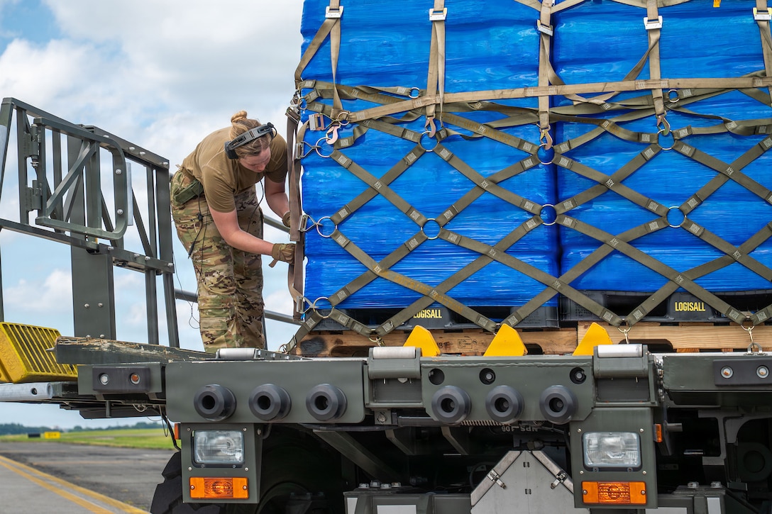 An airman wearing camouflage pants, gloves, a brown T-shirt and a communication headset adjusts straps on palleted bundles loaded on the back of a military truck.