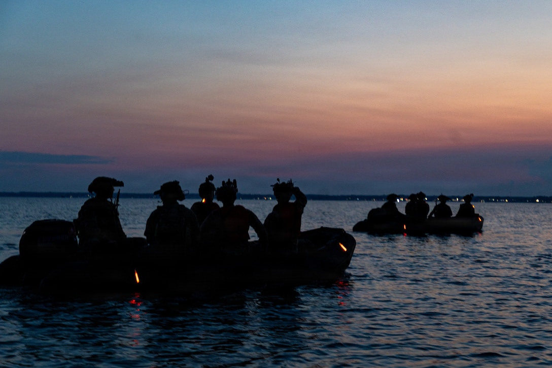 Two small tactical inflatable boats each carry five service members wearing helmets with affixed night-vision devices across a body of water under low light conditions.