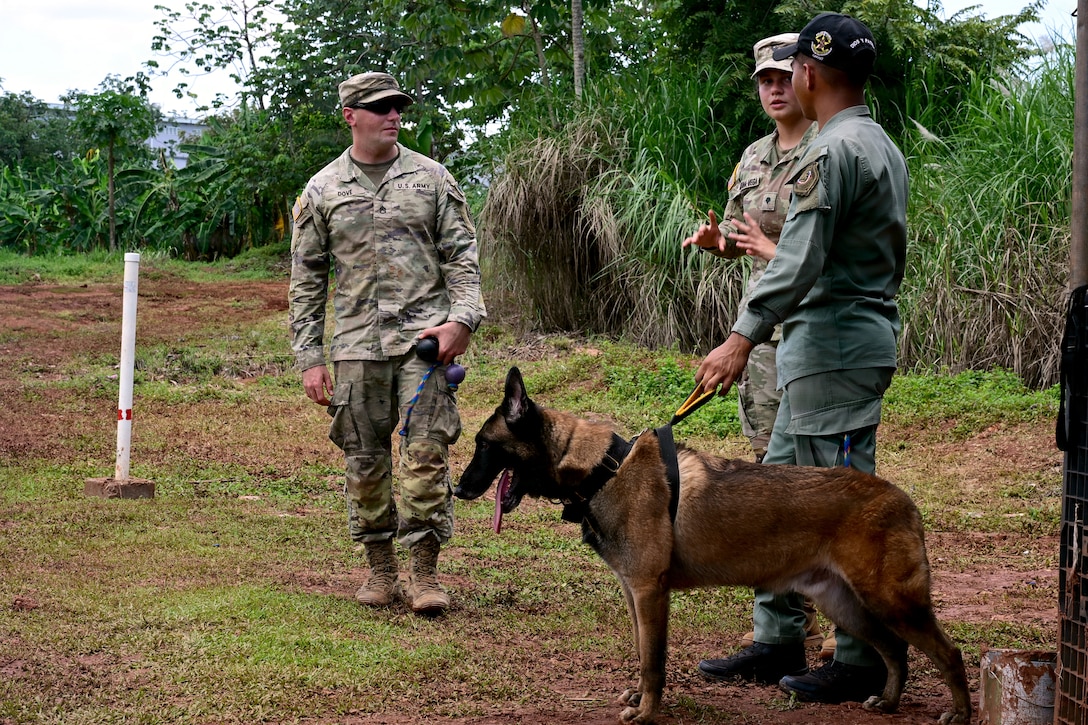 Two soldiers in camouflage uniforms speak with a man wearing blue fatigues holding a working dog on a leash during daytime with tropical foliage in the background.
