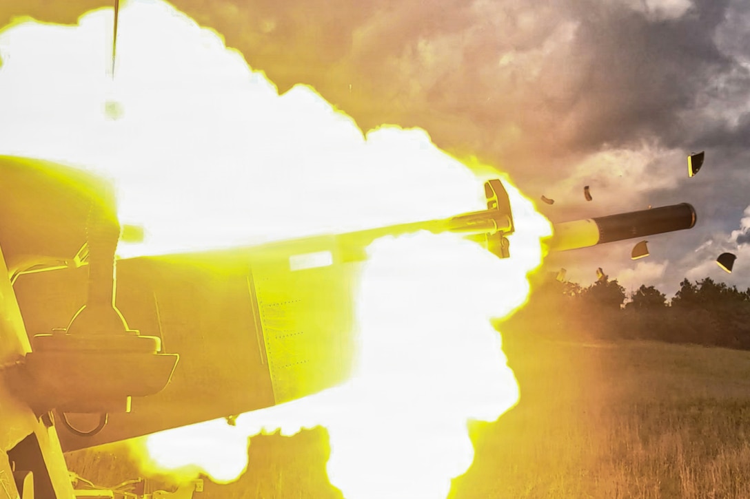 A rocket is fired from a launcher in a grassy field, leaving behind an orange blast on a cloudy day.