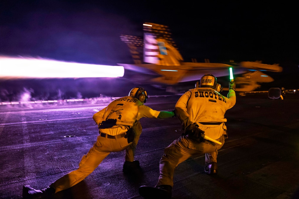 Two sailors wearing yellow flight-deck suits and headgear signal to a jet as it accelerates down a ship's runway in dark conditions, leaving a bright afterburner plume behind.