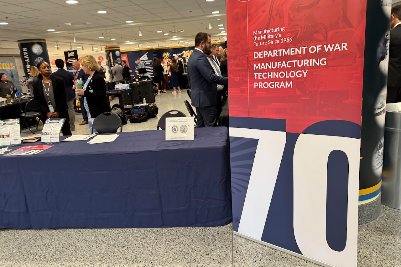 People in business attire view military technologies displayed on tables in a building.