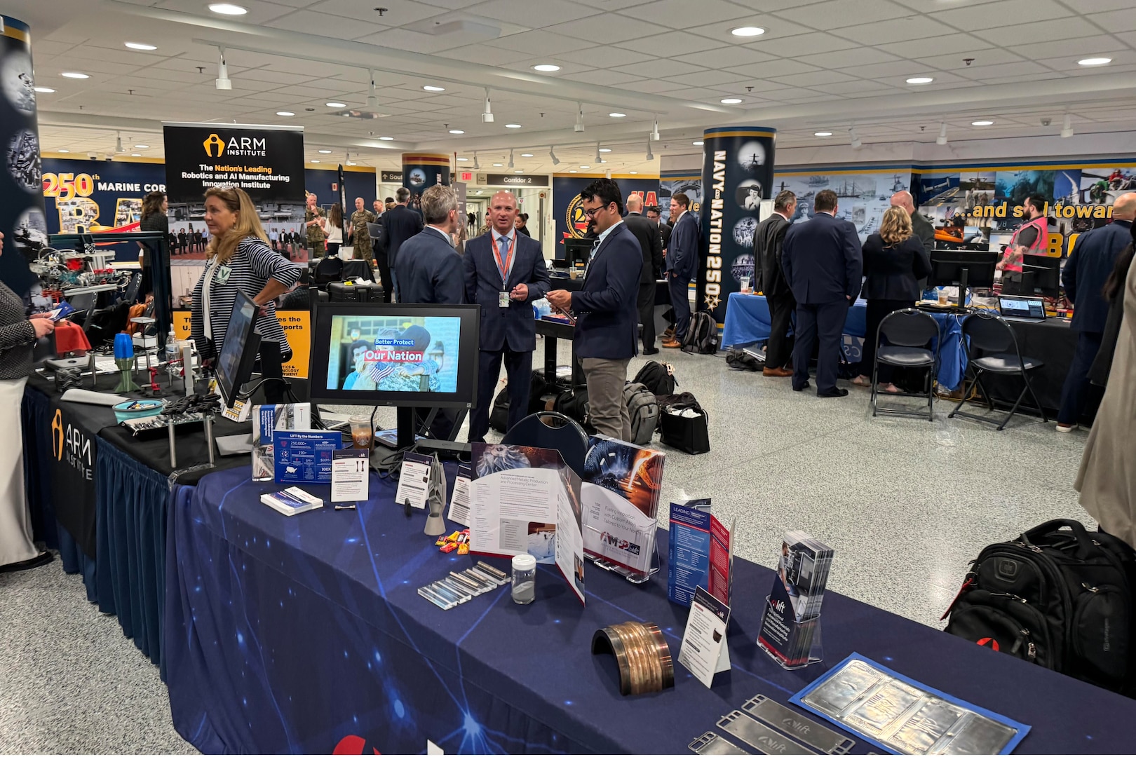People in business attire view military technologies displayed on tables in a building.