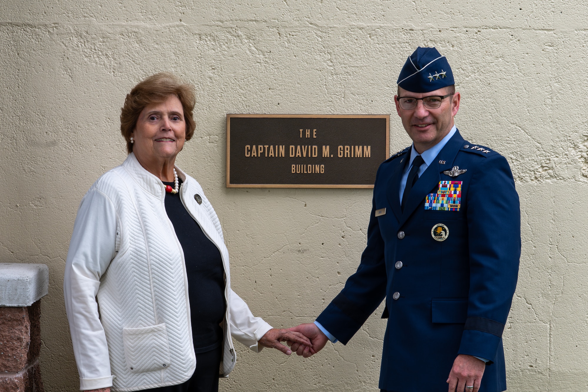Dana Grimm, wife of the late Capt. David M. Grimm, poses with U.S. Air Force Lt. Gen. Robert Davis, commander, Alaskan Command, Alaska North American Aerospace Defense Command Region, and Eleventh Air Force, in front of the newly dedicated Capt. David M. Grimm Building sign at Joint Base Elmendorf-Richardson, Alaska, Aug. 7, 2026.