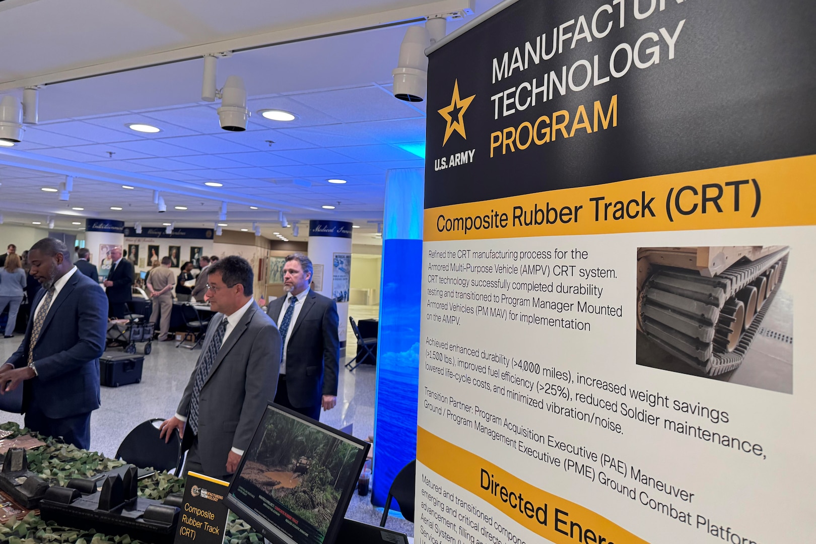 People in business attire view military technologies displayed on tables in a building.