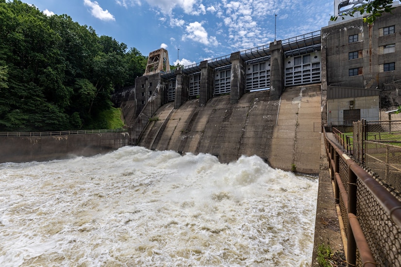 Raging water at the base of a dam in a wooded area and under a blue sky.