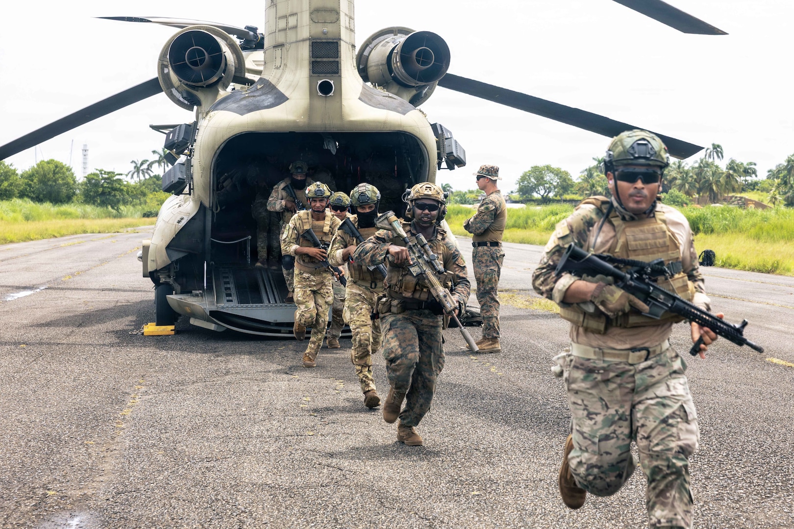 U.S. Marines and Panamanian service members in camouflage uniforms and carrying rifles run from the rear of a helicopter on a runway during daytime, as another person wearing a camouflage uniform observes.