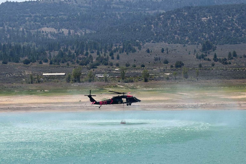 A helicopter flies low over a body of water collecting water in a bucket attached to the aircraft by a cable; trees and mountains are in the distance.