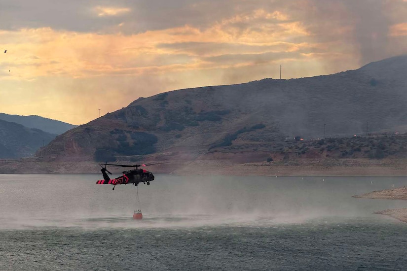 A helicopter flies low over a body of water collecting water in a bucket attached to the aircraft by a cable; the air is smoky and there are mountains in the distance.