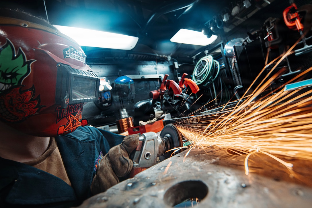 Sparks fly as a sailor wearing a protective face mask uses a tool to weld metal in a machine shop.
