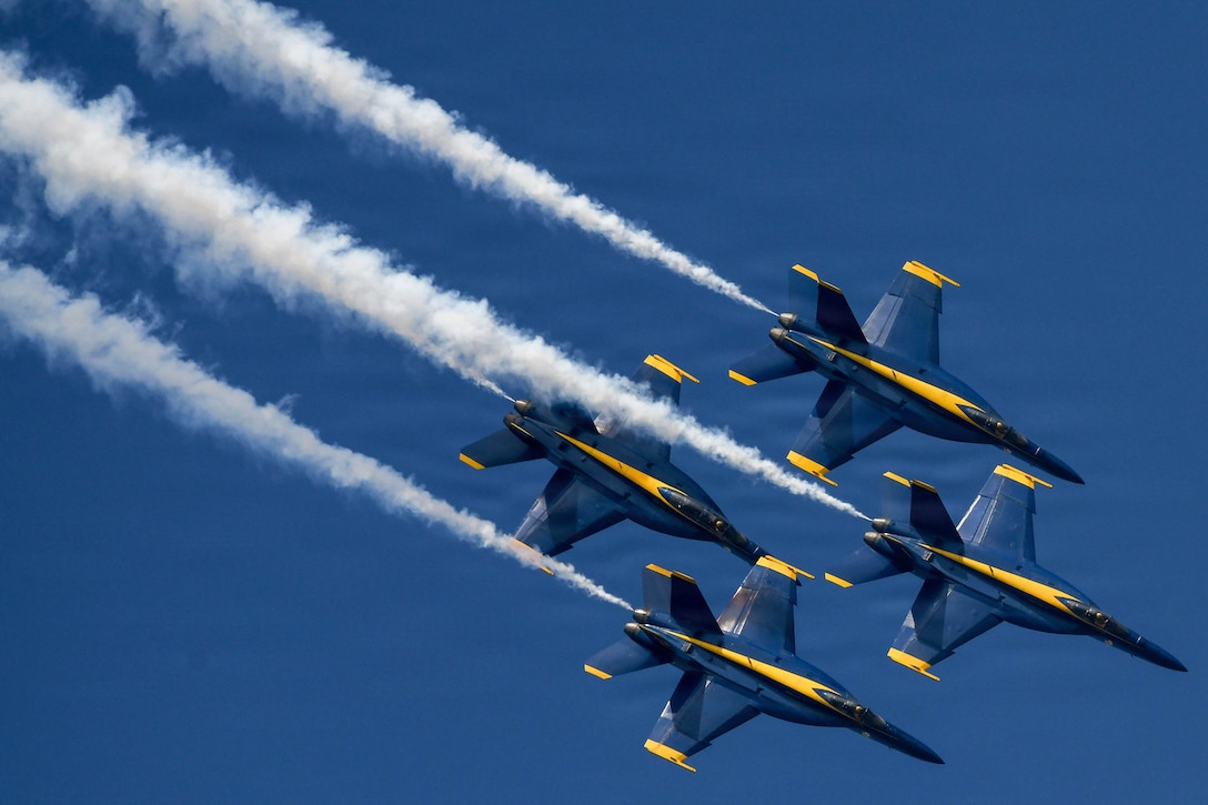 Four aircraft fly diagonally toward the bottom right in a blue sky, leaving behind trails of smoke.