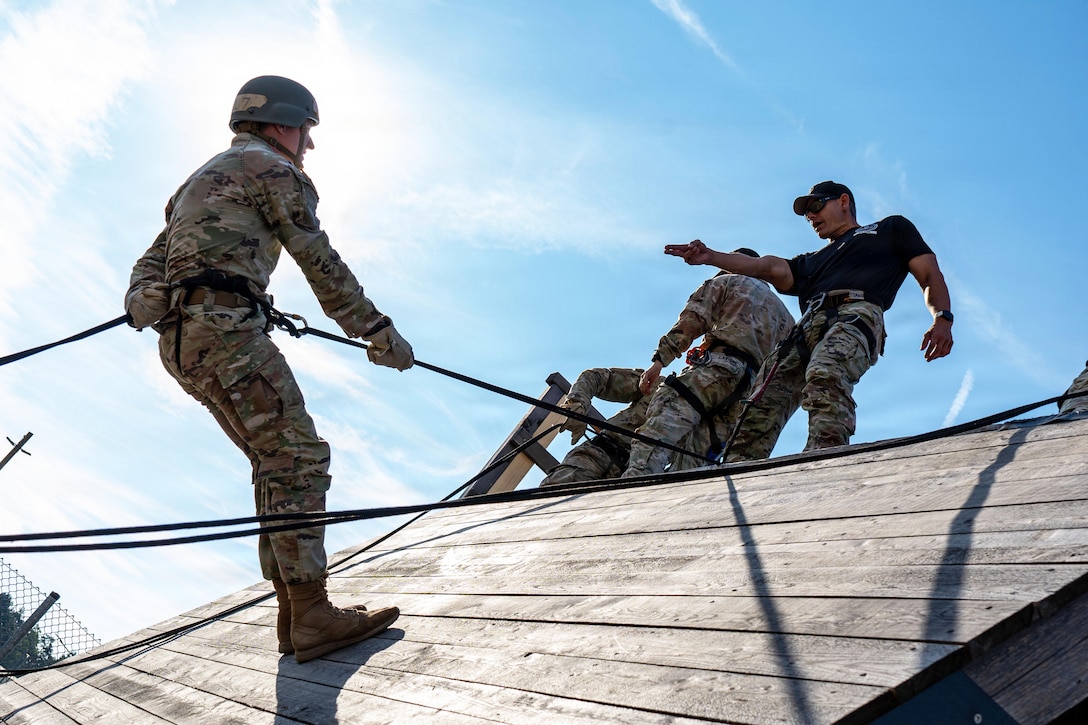 A soldier wearing a harness rappels down a wooden wall as an instructor points to them from above against a blue sky.