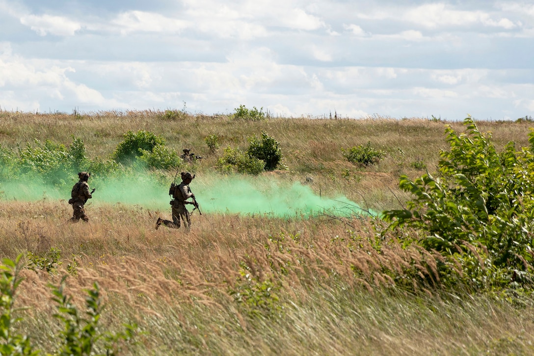 Two soldiers carrying weapons run through a field near green clouds of smoke.