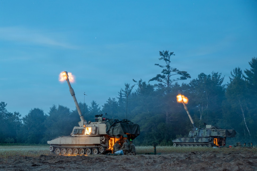 Flames shoot from two howitzers as they fire simultaneously against a deep blue sky, with soldiers manning them.