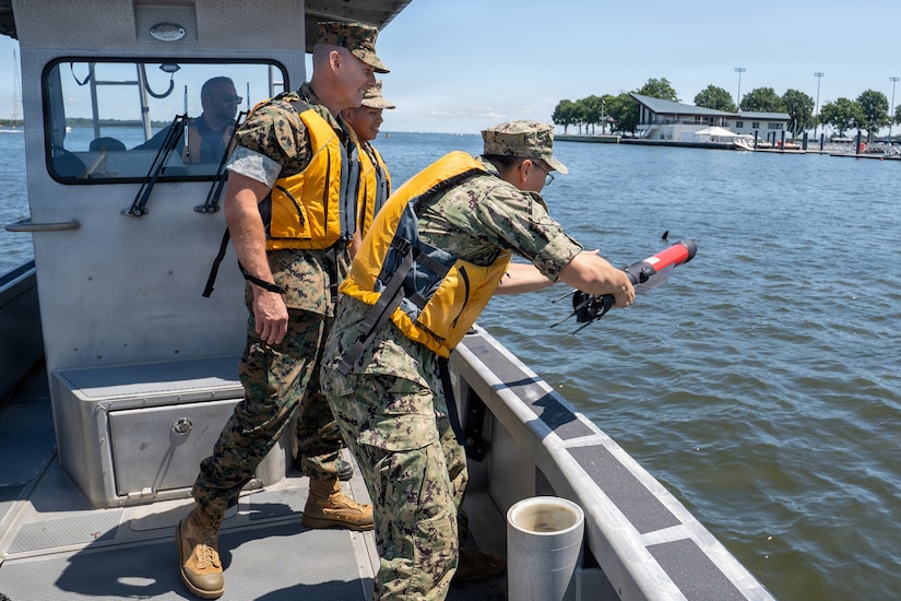 A man wearing a camouflage military uniform and life jacket throws an autonomous system into the water from a boat as three other people in similar attire observe.