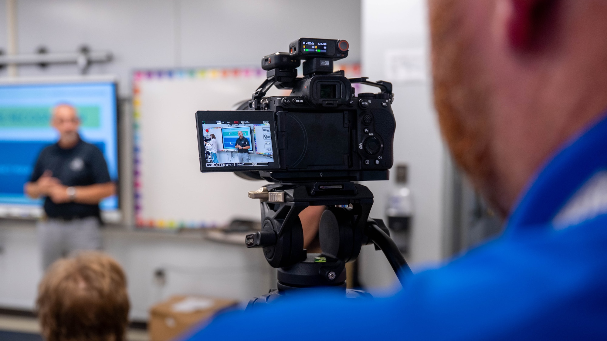 A photo of man in the foreground recording a man giving a lecture in the background.