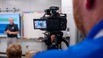 A photo of man in the foreground recording a man giving a lecture in the background.