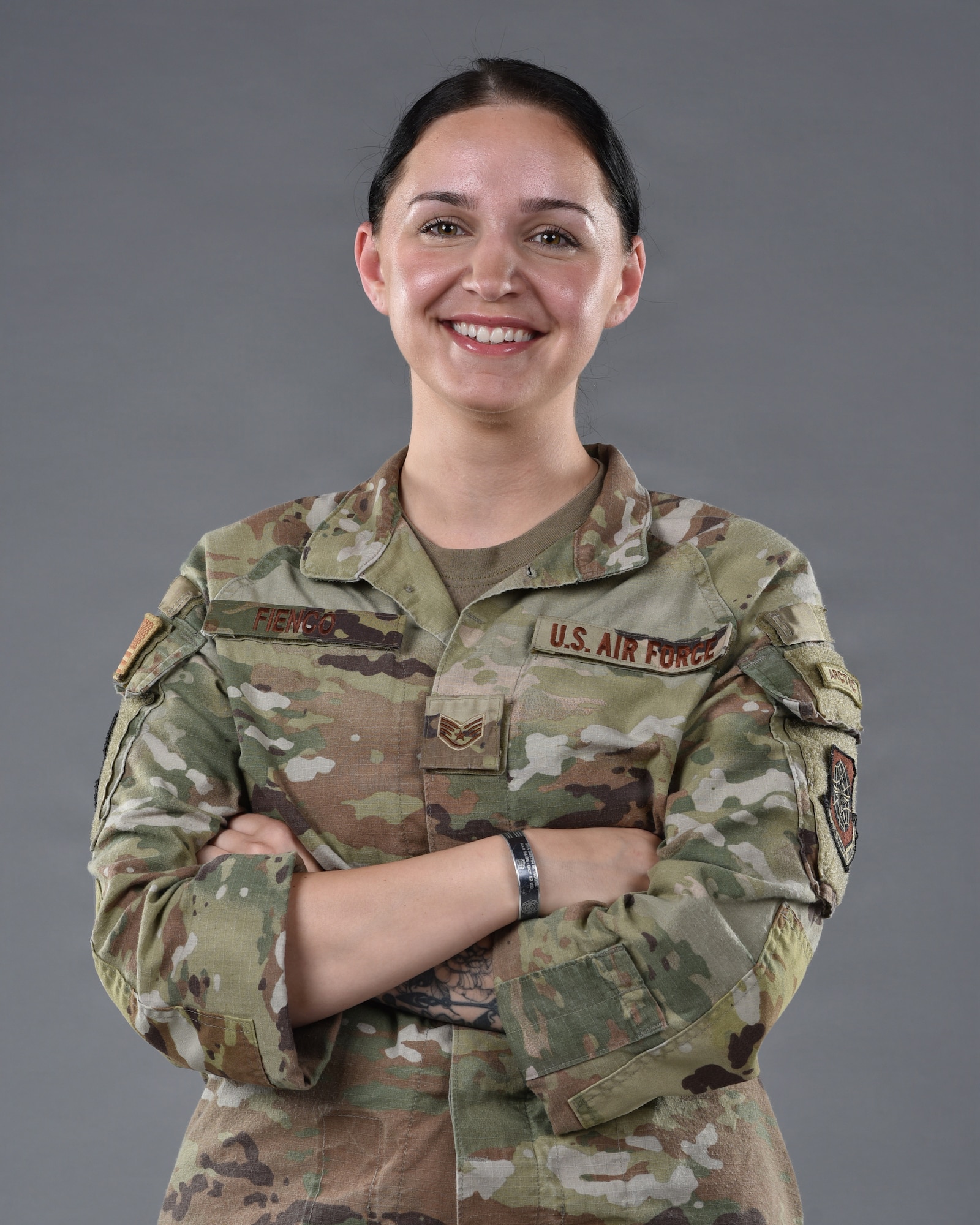 An Airman stands in front of a grey background with her arms crossed and smiles.