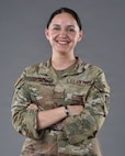 An Airman stands in front of a grey background with her arms crossed and smiles.