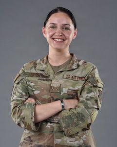 An Airman stands in front of a grey background with her arms crossed and smiles.