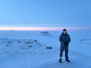 A service member stands atop a mountain with snow in the background.