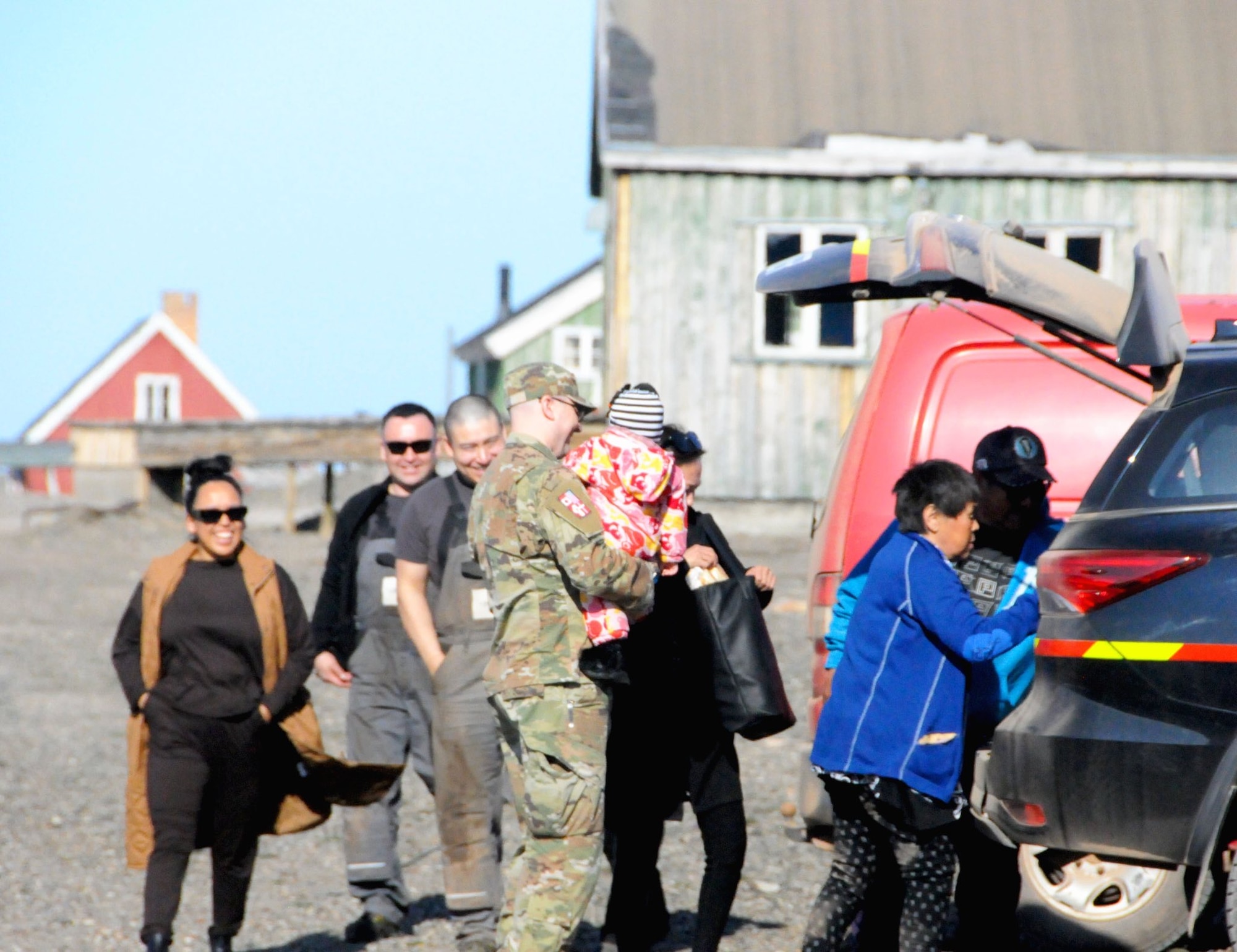 A military member holds a child by a car with other people surrounding them.