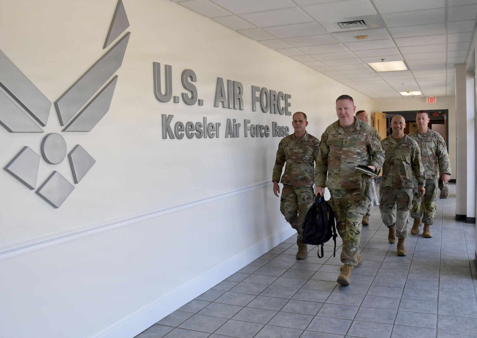 A group of men wearing U.S. Air Force uniforms walk down a hallway with words on the wall.