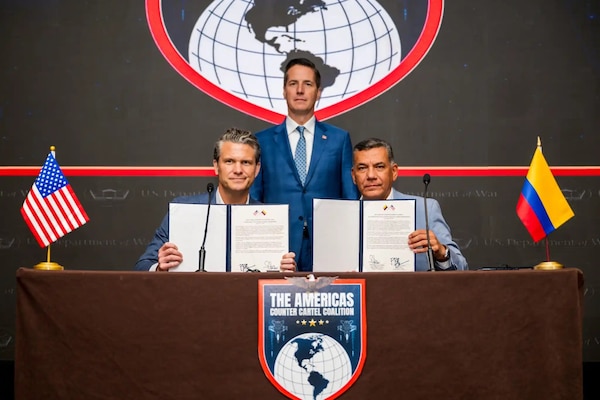 Two men in business attire are seated and holding up signed documents. One man stands behind them. On the table is a sign saying "The Americas Counter Cartel Coalition."