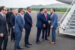 Secretary of War Pete Hegseth shakes hands with a civilian as a group greets him on a flight line at the base of aircraft steps.