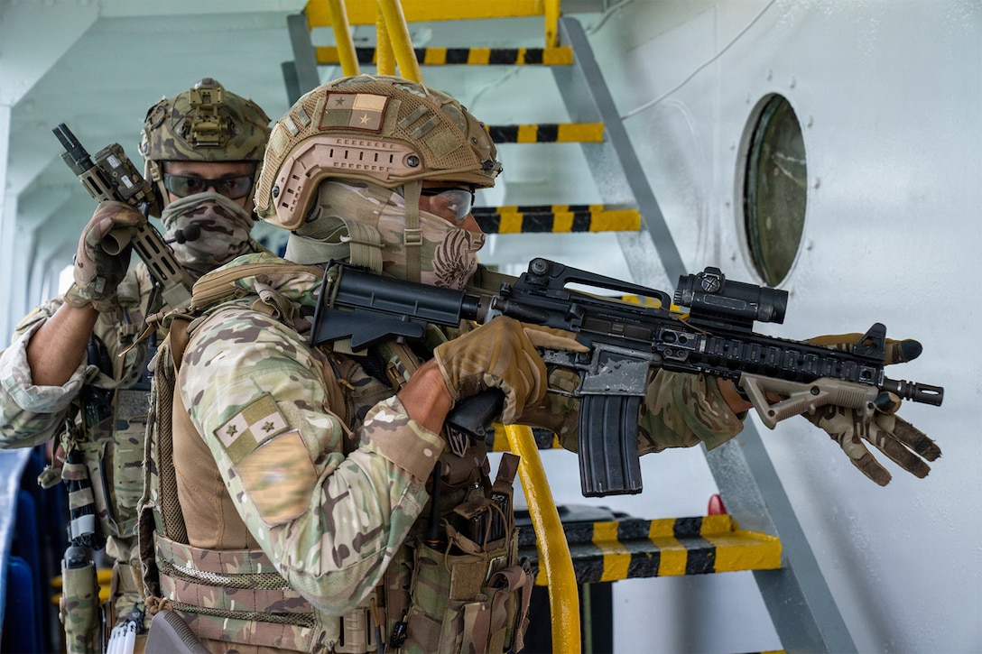 Two service members in camouflage uniforms hold rifles while moving near stairs on the deck of a ship.