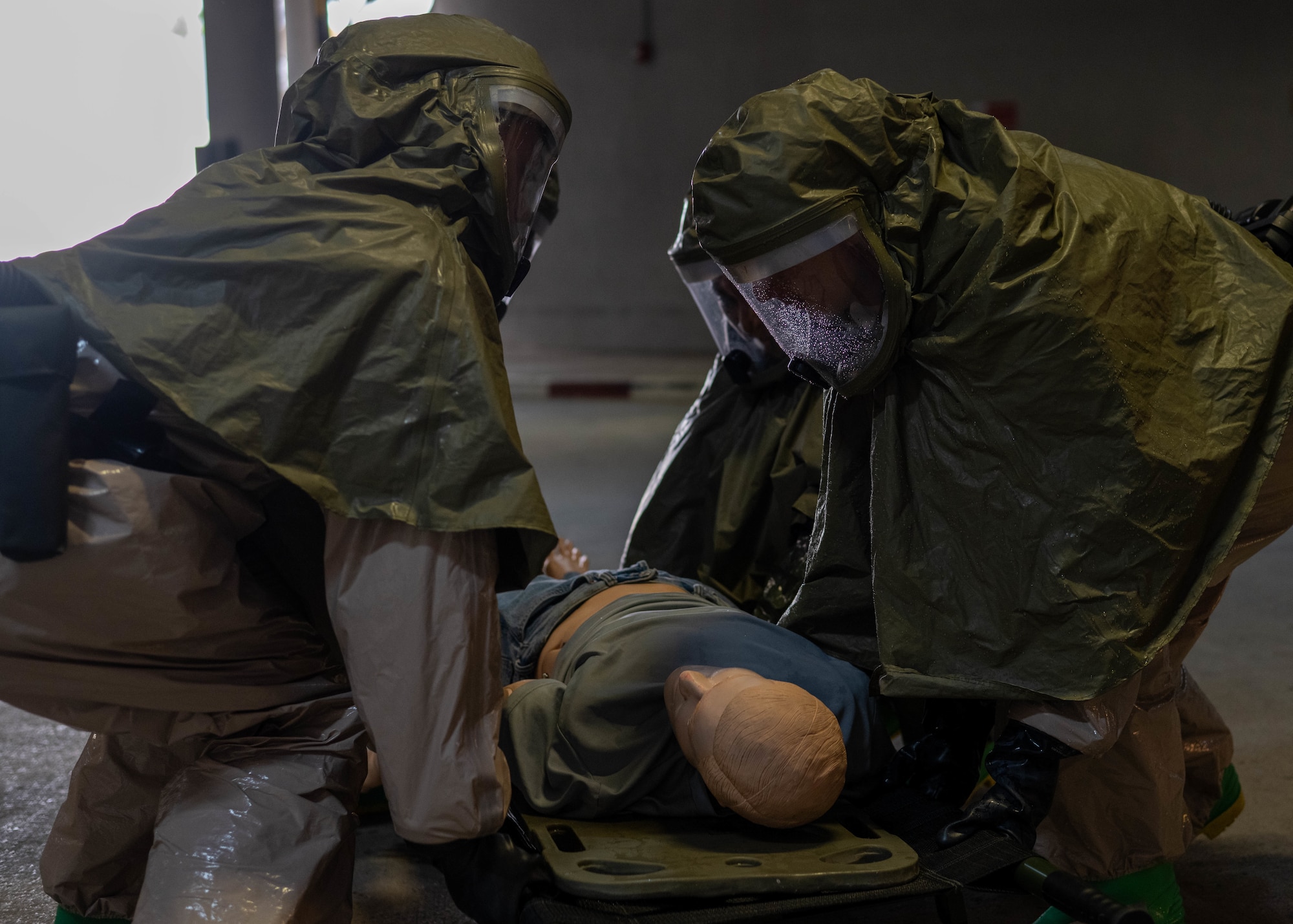 Four people in HAZMAT personal protective equipment, squat down to lift a weighted dummy on a liter carry.