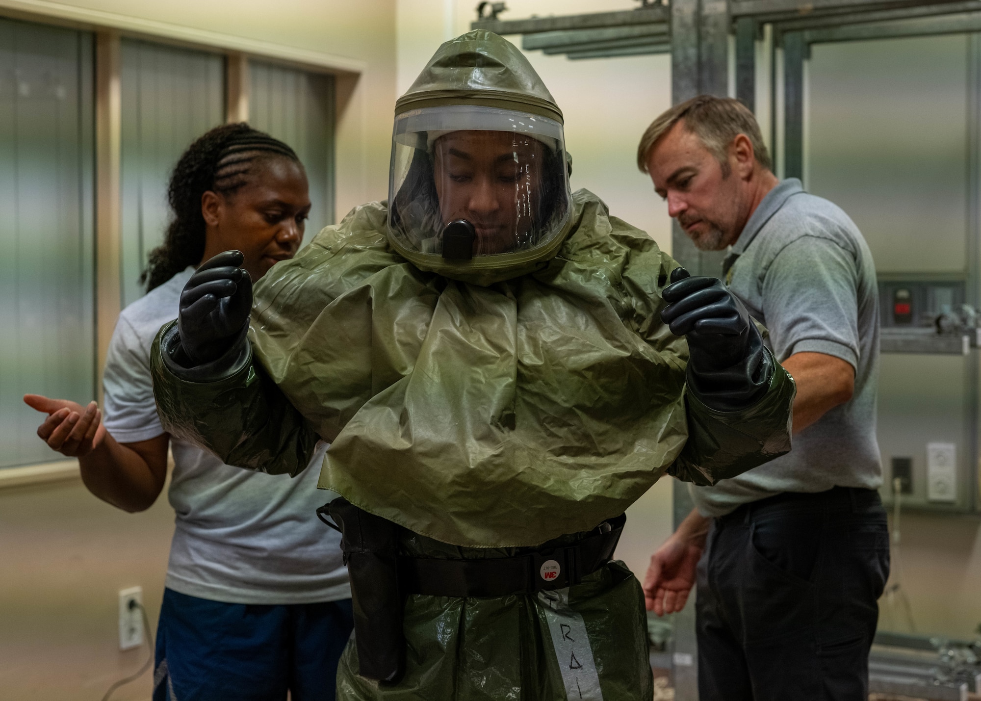 Airman in HAZMAT safety suit, stands in center frame as a man to her right and a woman to her left, assess her uniform to complete uniform checks.