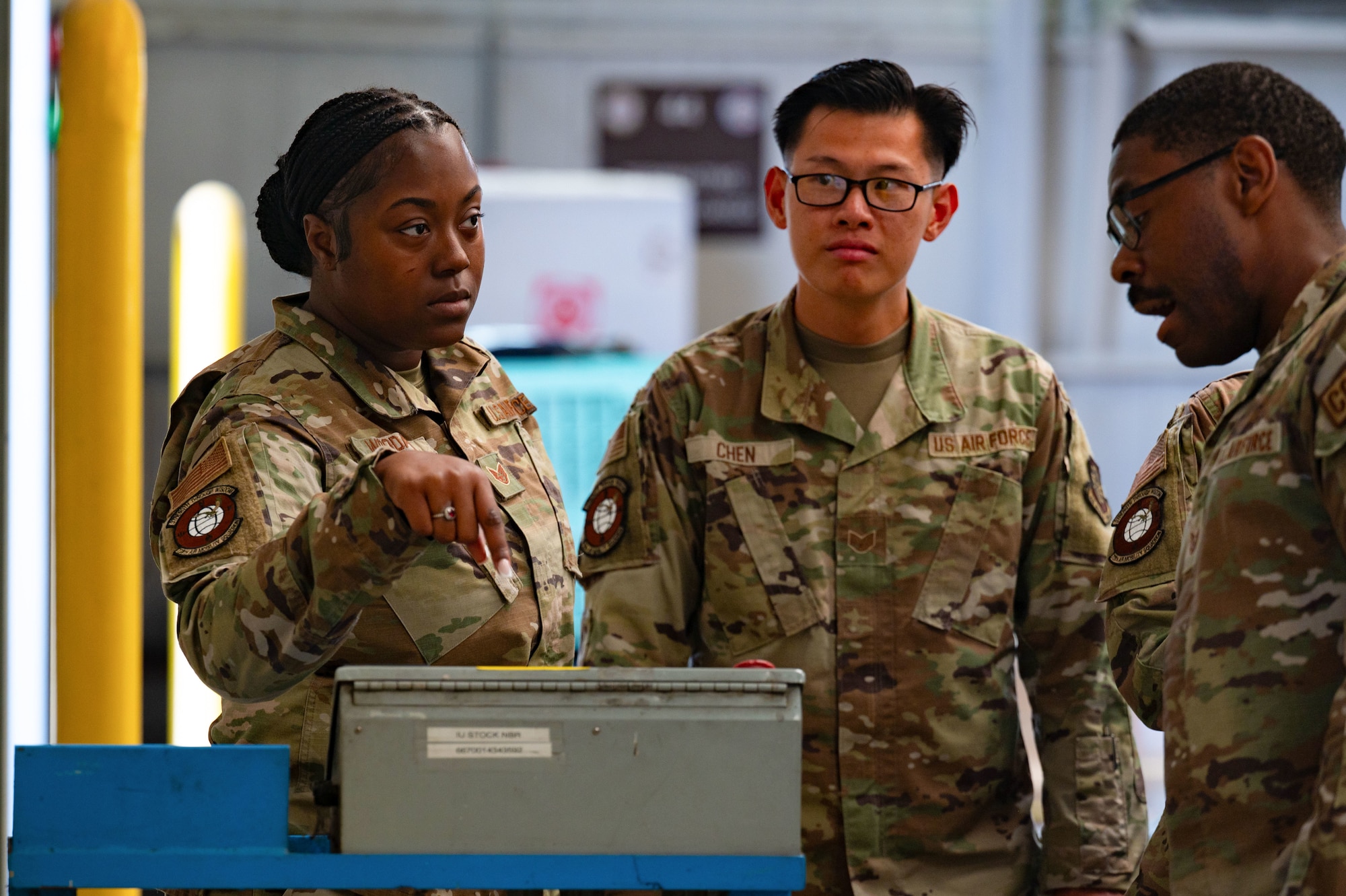 A group of airmen stand and talk as one points at a grey box.
