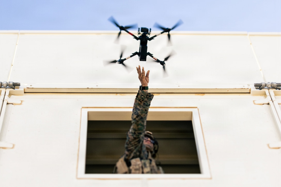 A Marine leans out an open window and extends their arm upward to release a drone, which hovers just above the Marine's hand.