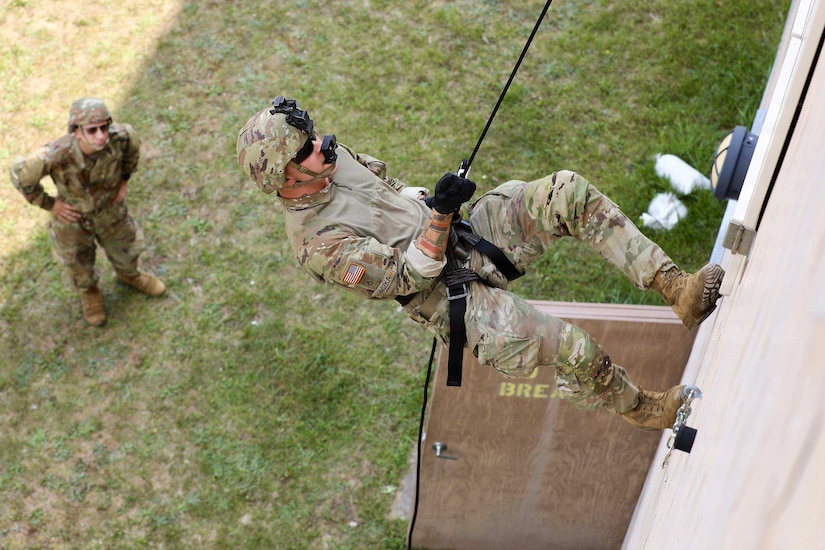 A man wearing a camouflage military uniform rappels down the front of a building as another man in similar attire observes on the ground.