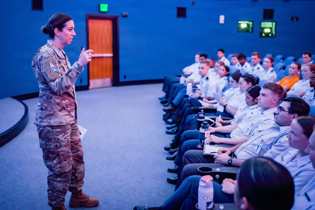 A woman wearing a camouflage military uniform stands at the front of an auditorium and speaks to people dressed in military dress uniforms who are seated.
