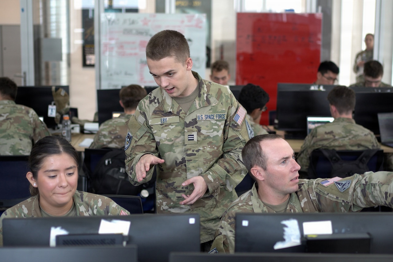 A man wearing a camouflage military uniform gestures at a computer screen that a woman wearing similar attire is seated in front of; another man also in similar attire and seated points in the opposite direction.