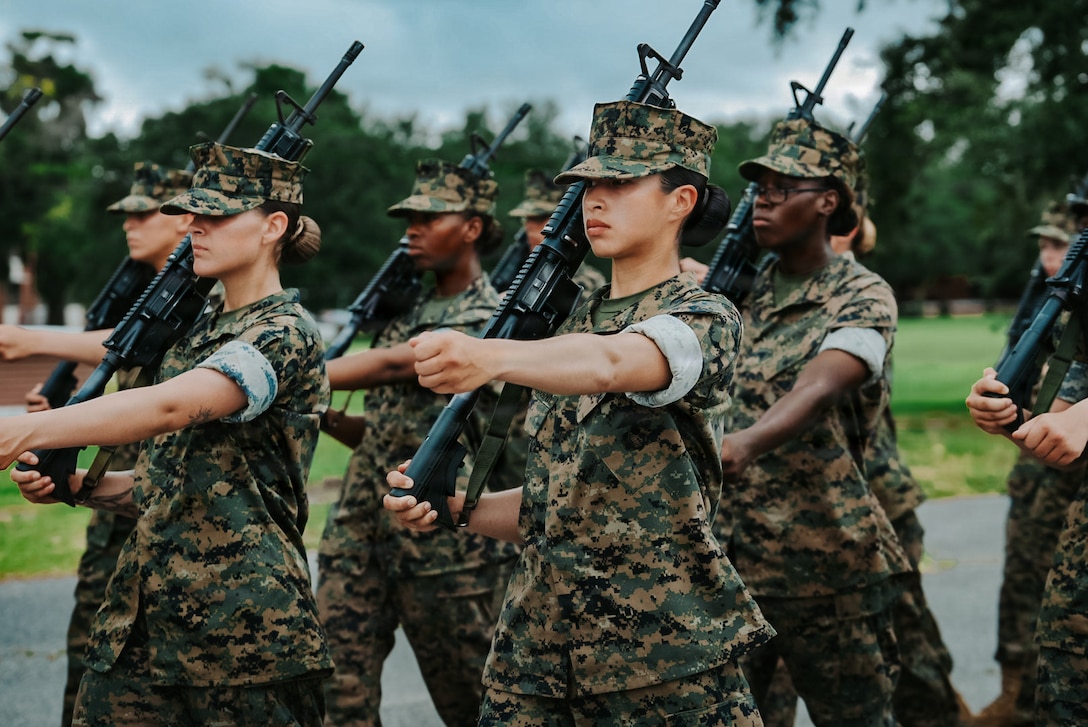 Marine Corps recruits in camouflage uniforms and holding rifles extend their left arms forward while marching in formation during daytime.