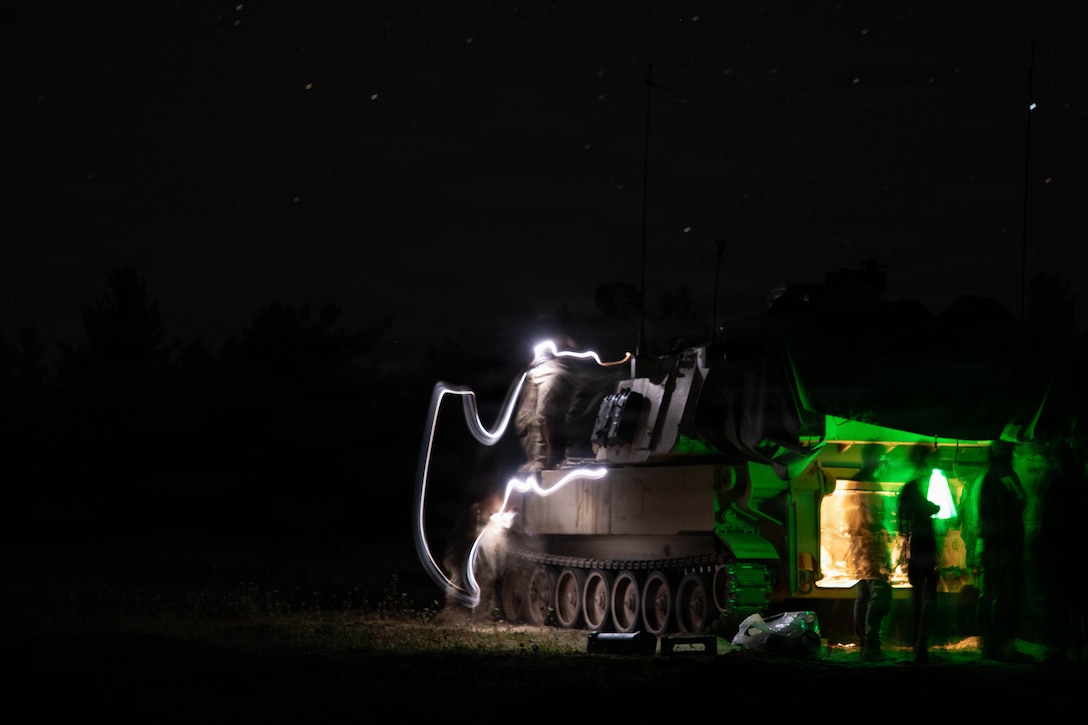Three soldiers stand near a military vehicle in dark conditions, illuminated by green light; streaks of white light are at the vehicle's front, with a person standing above its treads.
