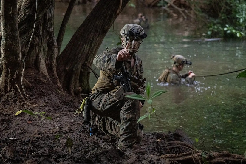 In a jungle environment, a man in a camouflage military uniform kneels on a riverbank as another man in similar attire stands in the water.