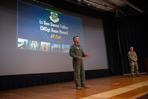 A white man in a flight suit stands in the center of a stage with another man in a multi-cam uniform to his right. A slide show presentation reading "Lt Gen Daniel Tulley and CMSgt Raun Howard, All Call" behind them.