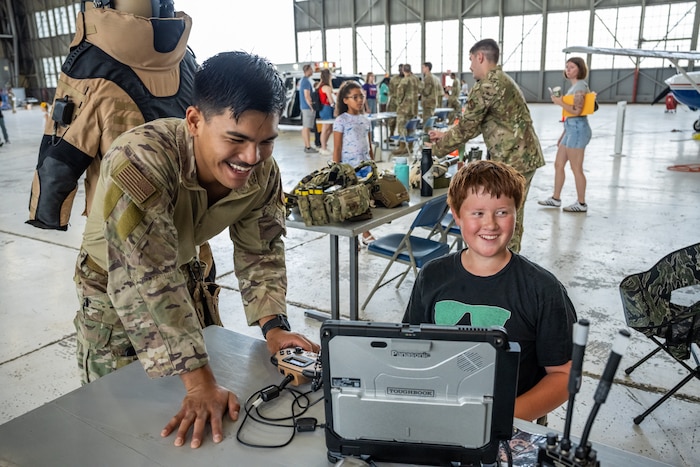 A man, wearing camouflage military uniform, laughs alongside a young boy sitting behind a tactical laptop