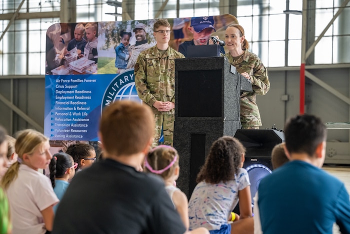 A woman, wearing camouflage military uniform, stands behind a podium and microphone in front of a group of children