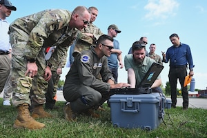 Men in Air Force uniforms and business attire stand in field on sunny day, looking at a black laptop
