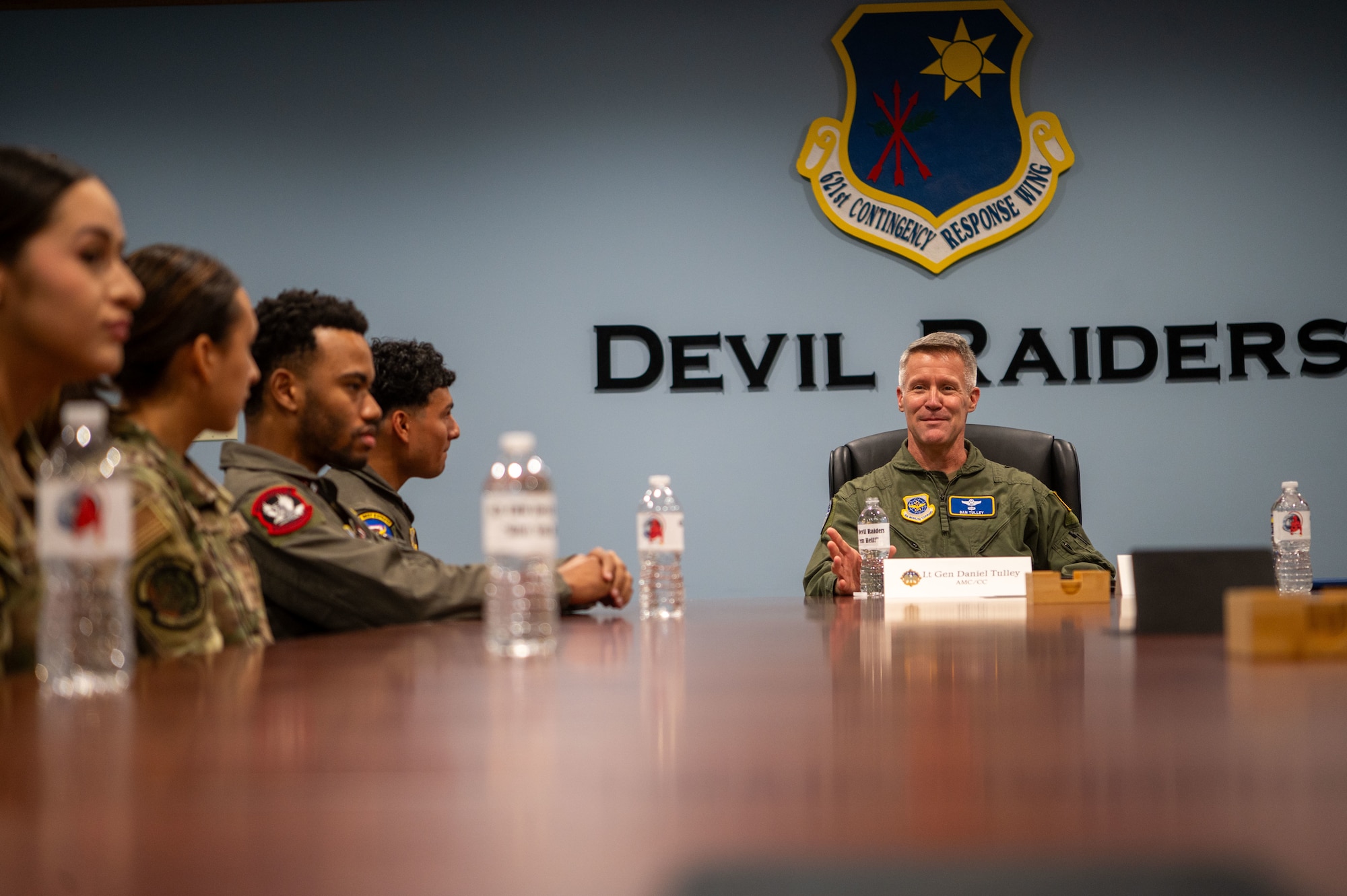 A white man in a flight suit sits at the top of a conference table in the top right of the photo with men and women in uniform sitting to his left, with the words "Devil Raiders" on the wall behind them.
