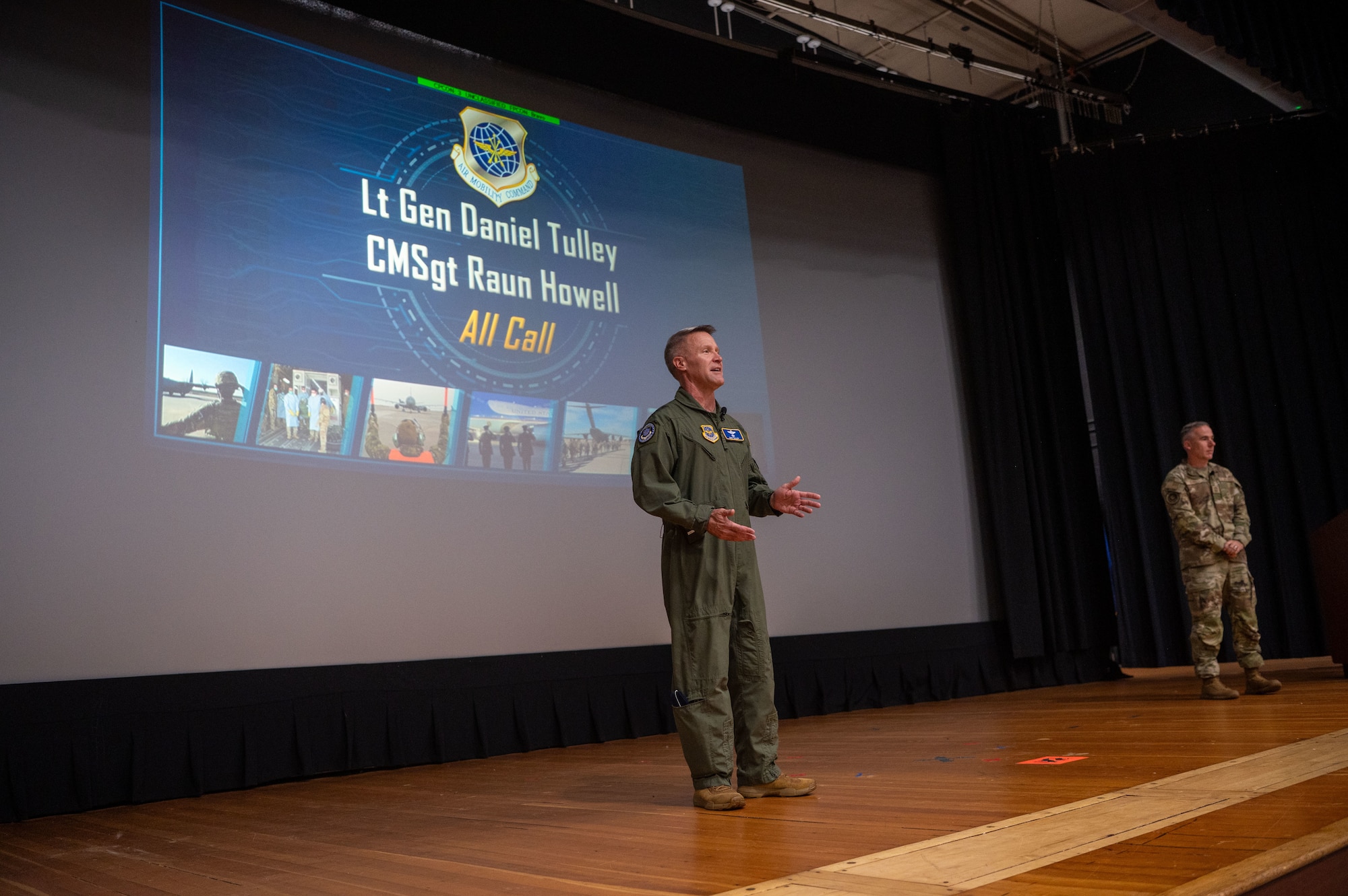 A white man in a flight suit stands in the center of a stage with another man in a multi-cam uniform to his right. A slide show presentation reading "Lt Gen Daniel Tulley and CMSgt Raun Howard, All Call" behind them.